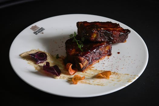 Travers de porc laqués de sauce BBQ, présentés sur une assiette avec garnitures colorées et sauce.