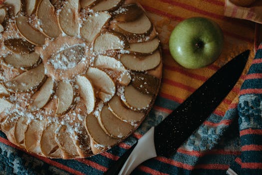 Tarte aux pommes maison décorée de tranches de pommes, avec une pomme verte et un couteau sur un torchon coloré.