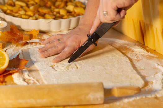 Main coupant une pâte à tarte sur un plan de travail fariné, avec des pommes en dés en arrière-plan.