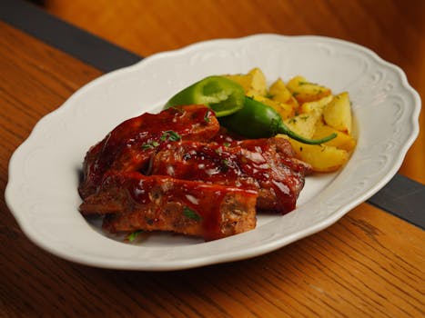 Travers de porc nappés de sauce BBQ, accompagnés de pommes de terre rôties et de piments sur une assiette blanche.
